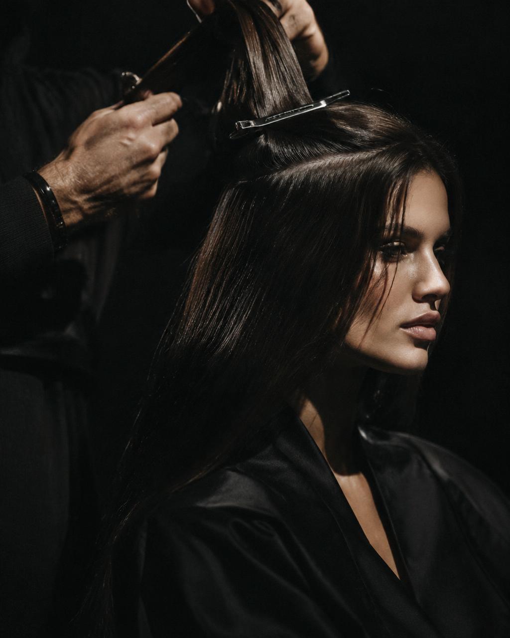 A stylist sectioning long dark hair with a metal clip, lit against black